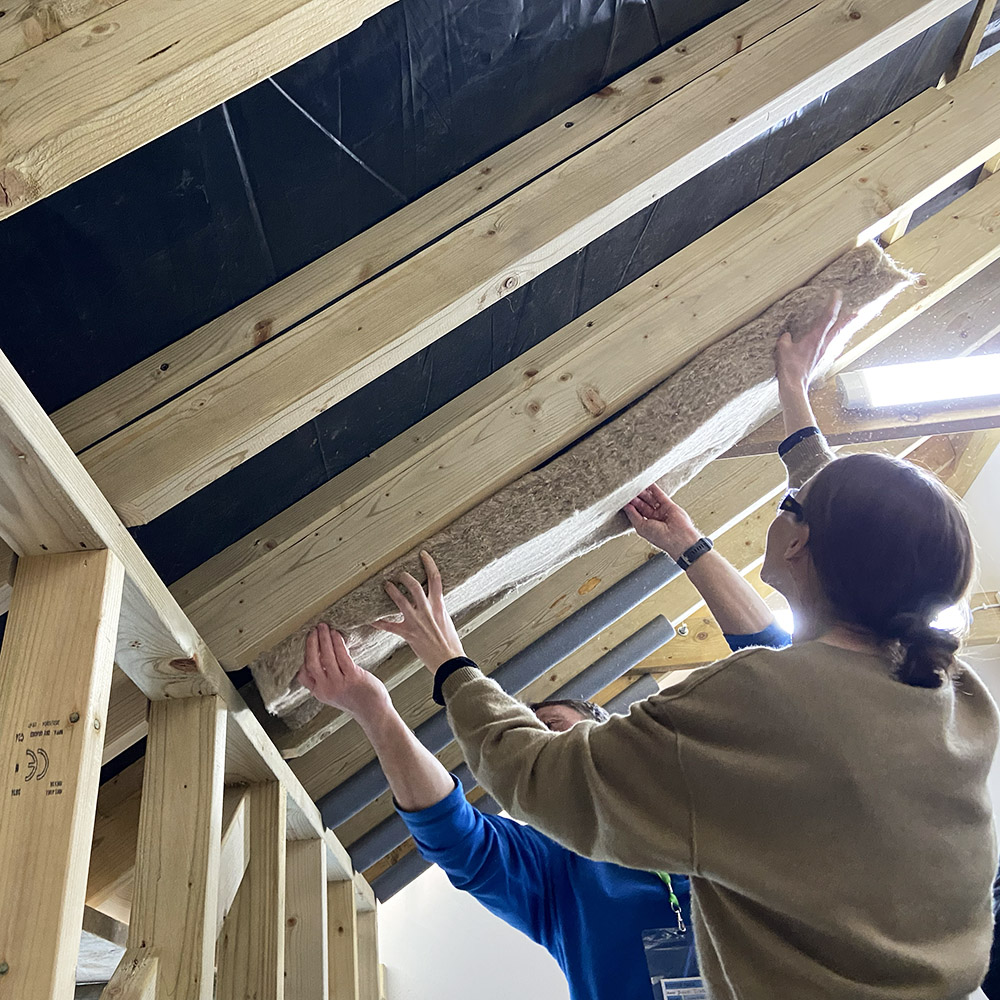 Two people installing hemp insulation between rafters to underside of pitched roof