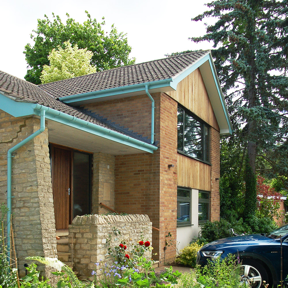 Modernist house built with stone, brick, and timber cladding, within setting of mature trees.