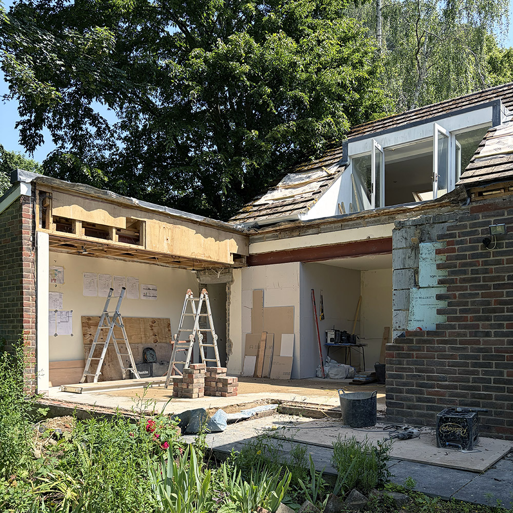 Corner of 1970s brick house with roof removed and under construction, with trees and blue skies above
